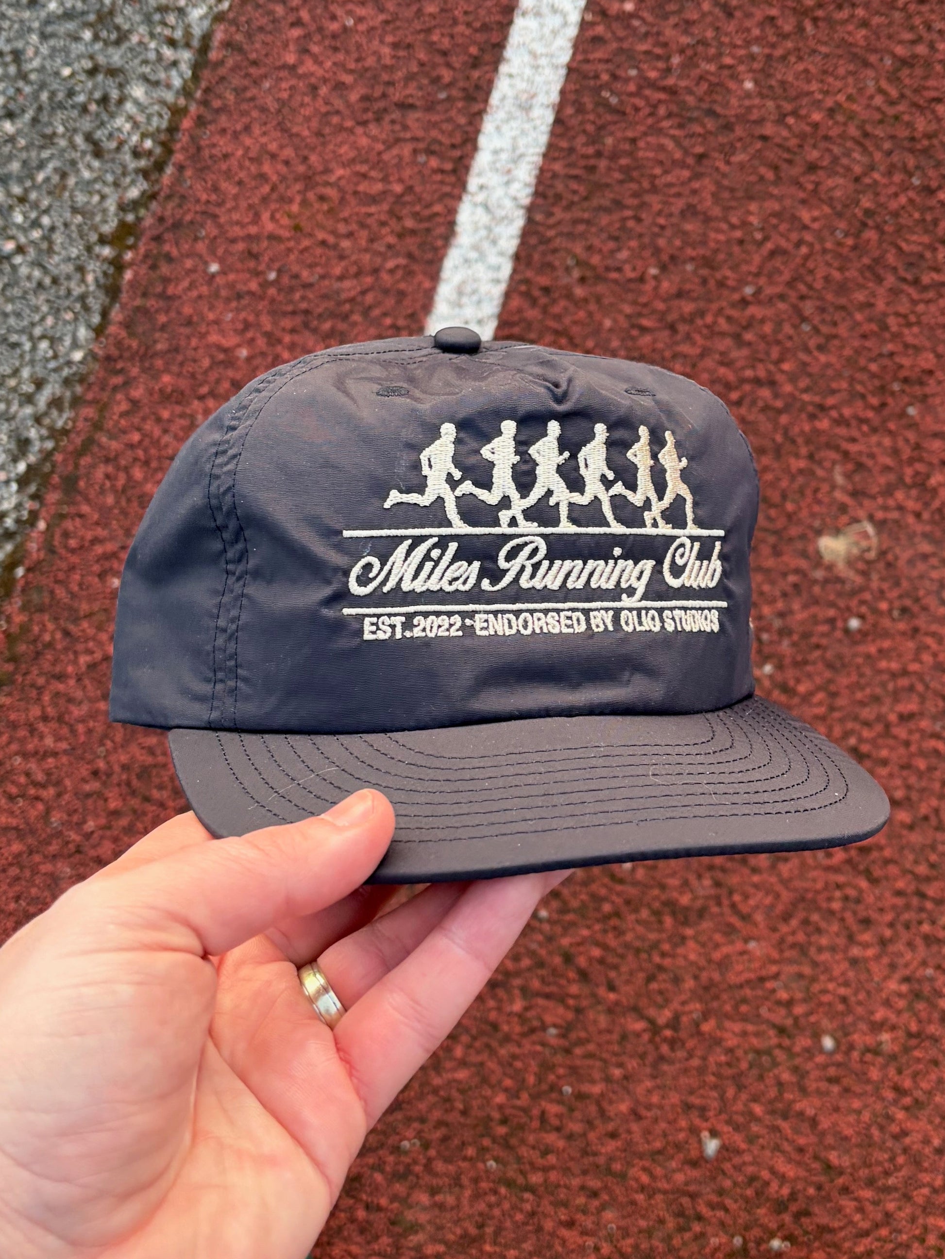 Navy blue cap with 'Miles Running Club' logo held above a running track